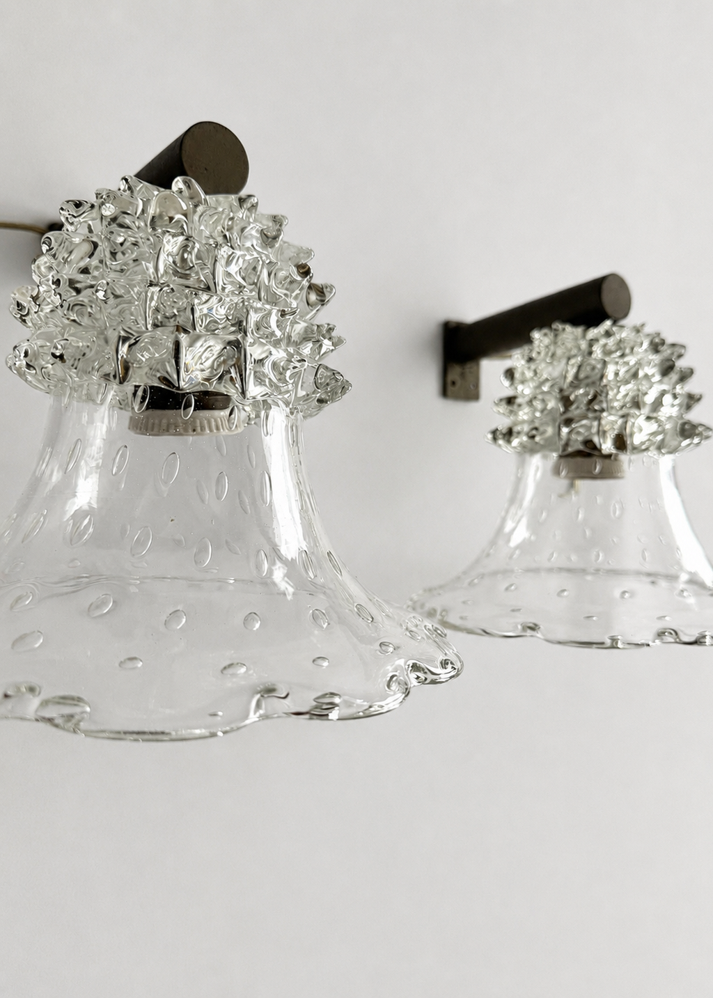 Two clear glass bell-shaped wall sconces with textured silver floral caps and dark metal wall mounts, shown against a plain light background.