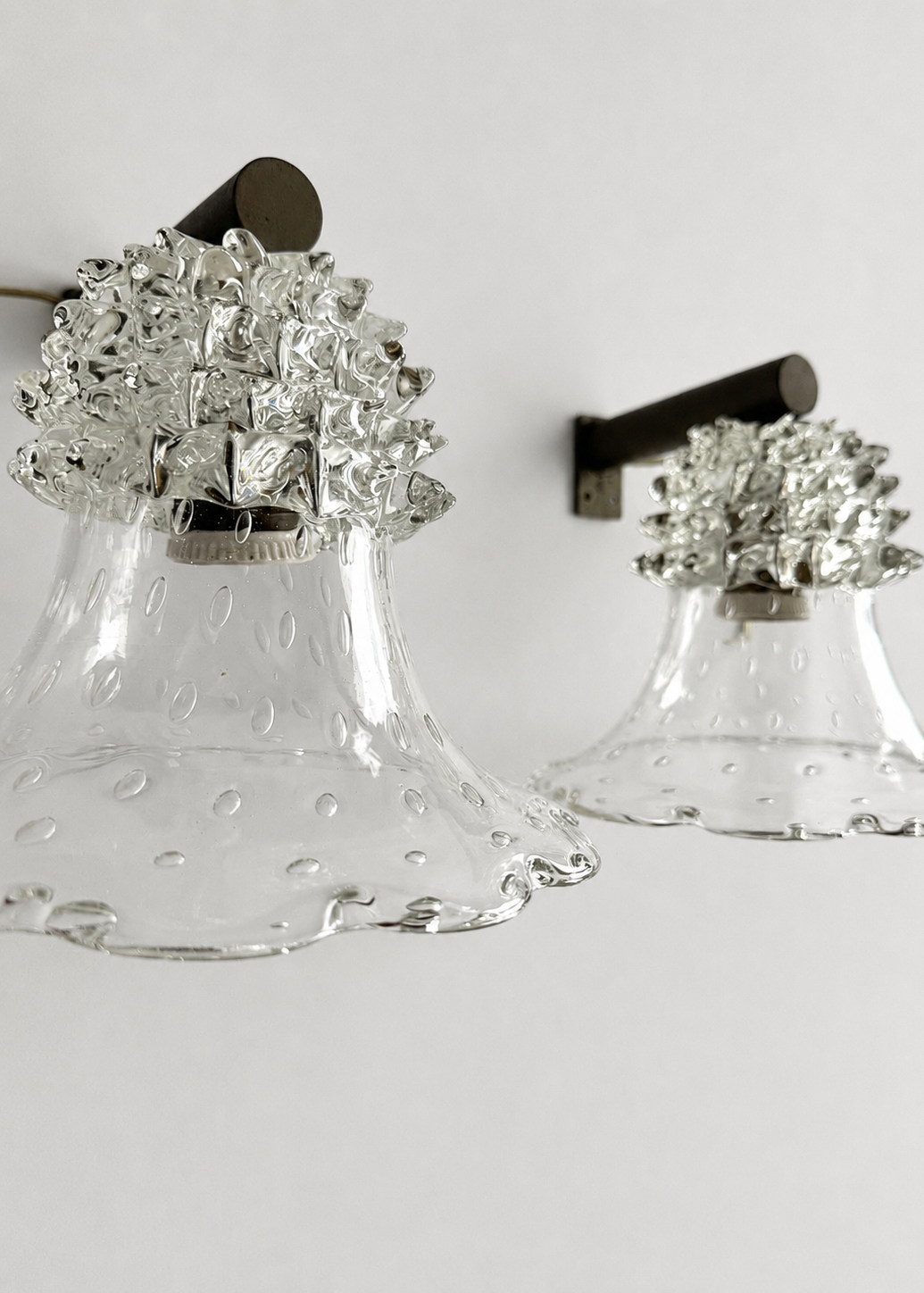 Two clear glass bell-shaped wall sconces with textured silver floral caps and dark metal wall mounts, shown against a plain light background.