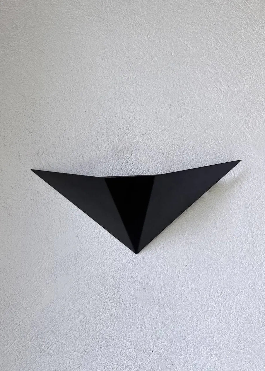 Two black geometric triangular wall-mounted light fixtures or decorative panels placed on a concrete floor against a white textured wall.