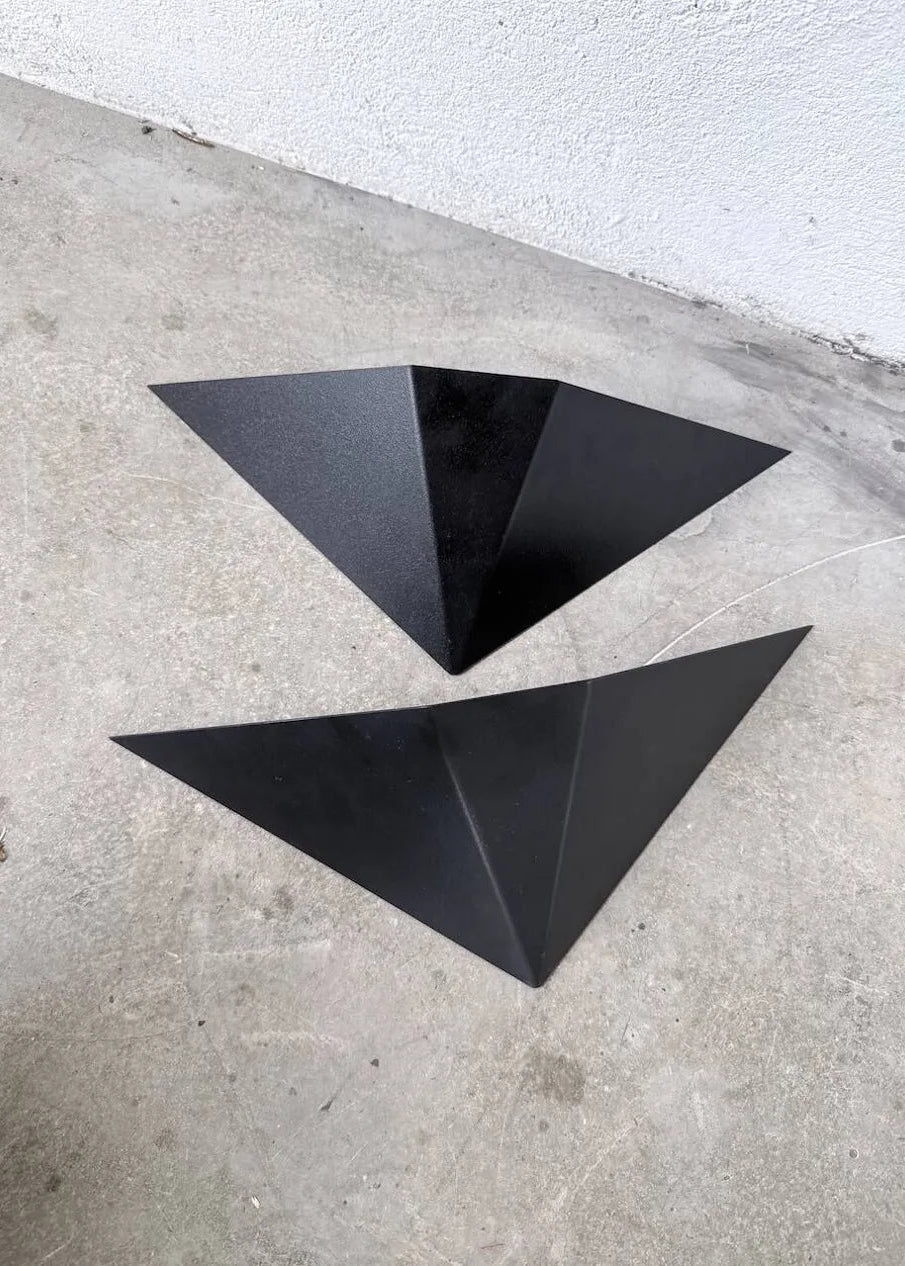 Two black geometric triangular wall-mounted light fixtures or decorative panels placed on a concrete floor against a white textured wall.