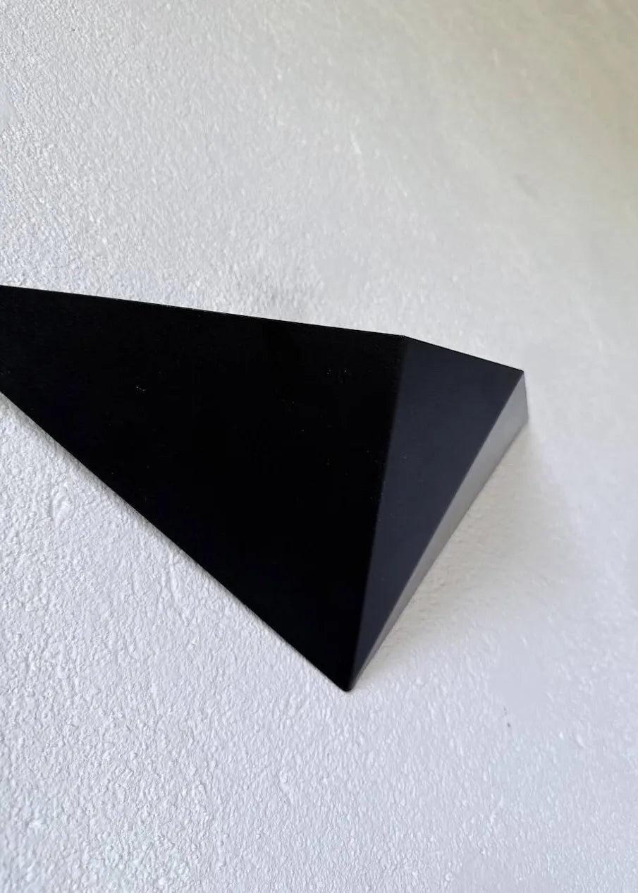 Two black geometric triangular wall-mounted light fixtures or decorative panels placed on a concrete floor against a white textured wall.