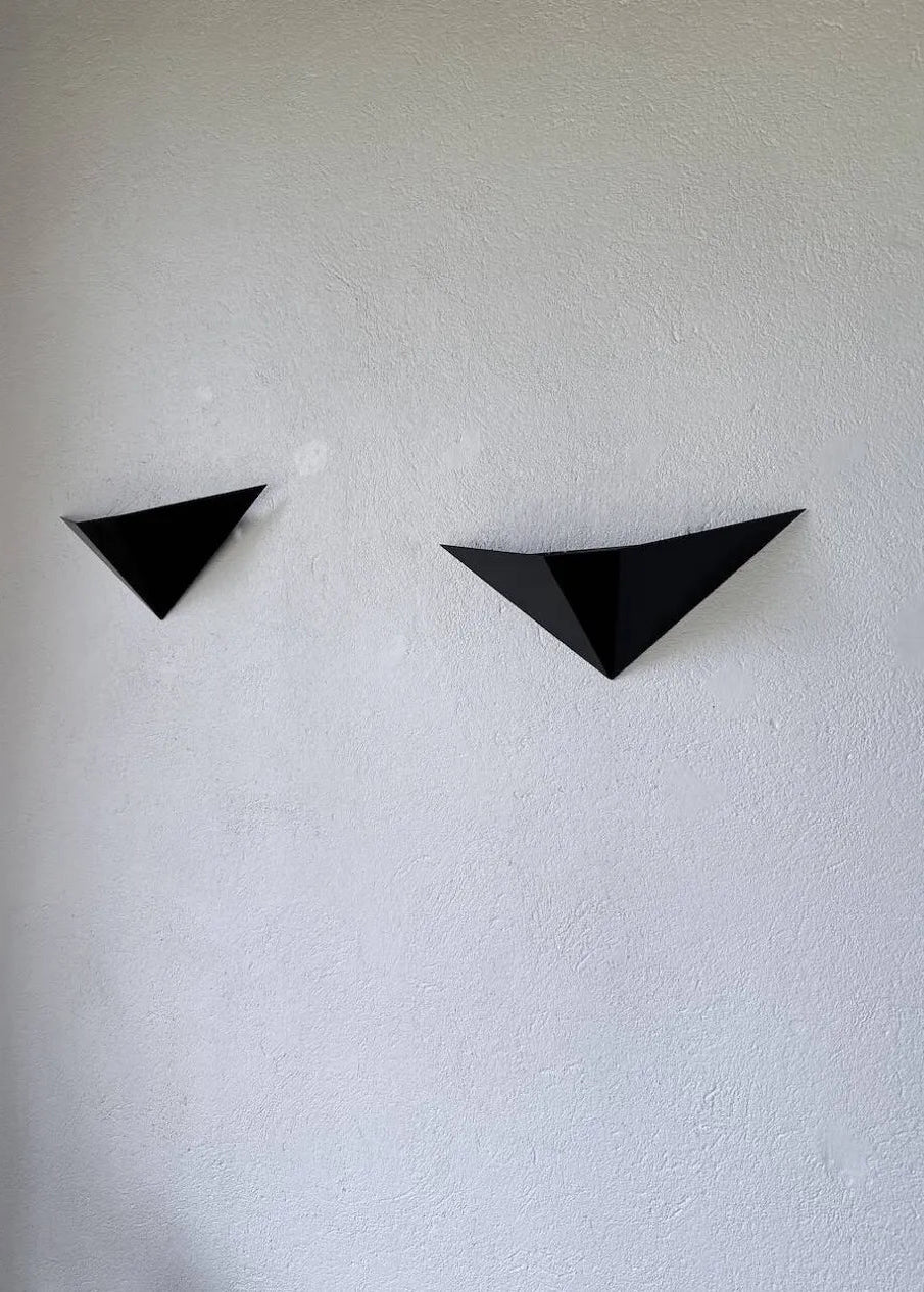 Two black geometric triangular wall-mounted light fixtures or decorative panels placed on a concrete floor against a white textured wall.