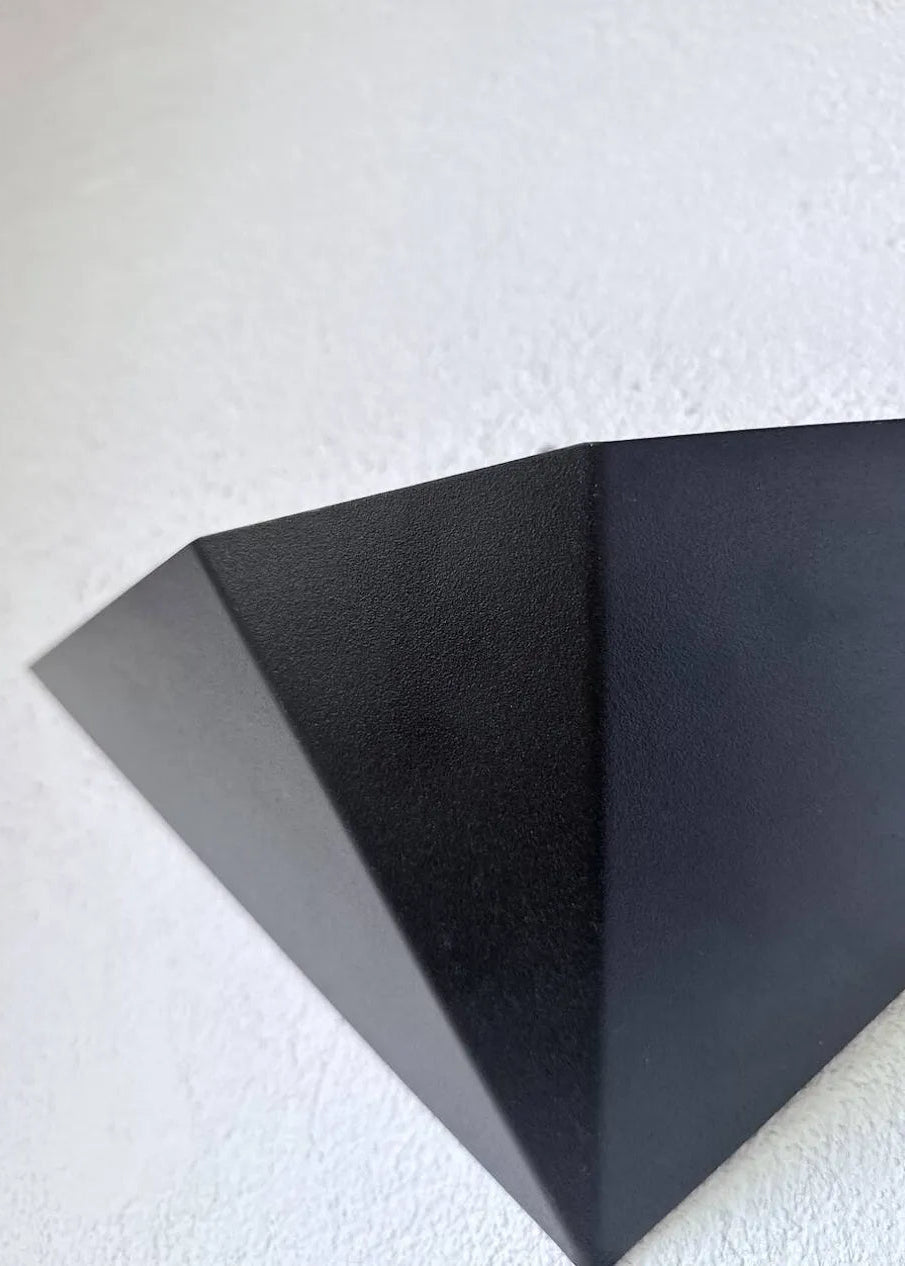 Two black geometric triangular wall-mounted light fixtures or decorative panels placed on a concrete floor against a white textured wall.