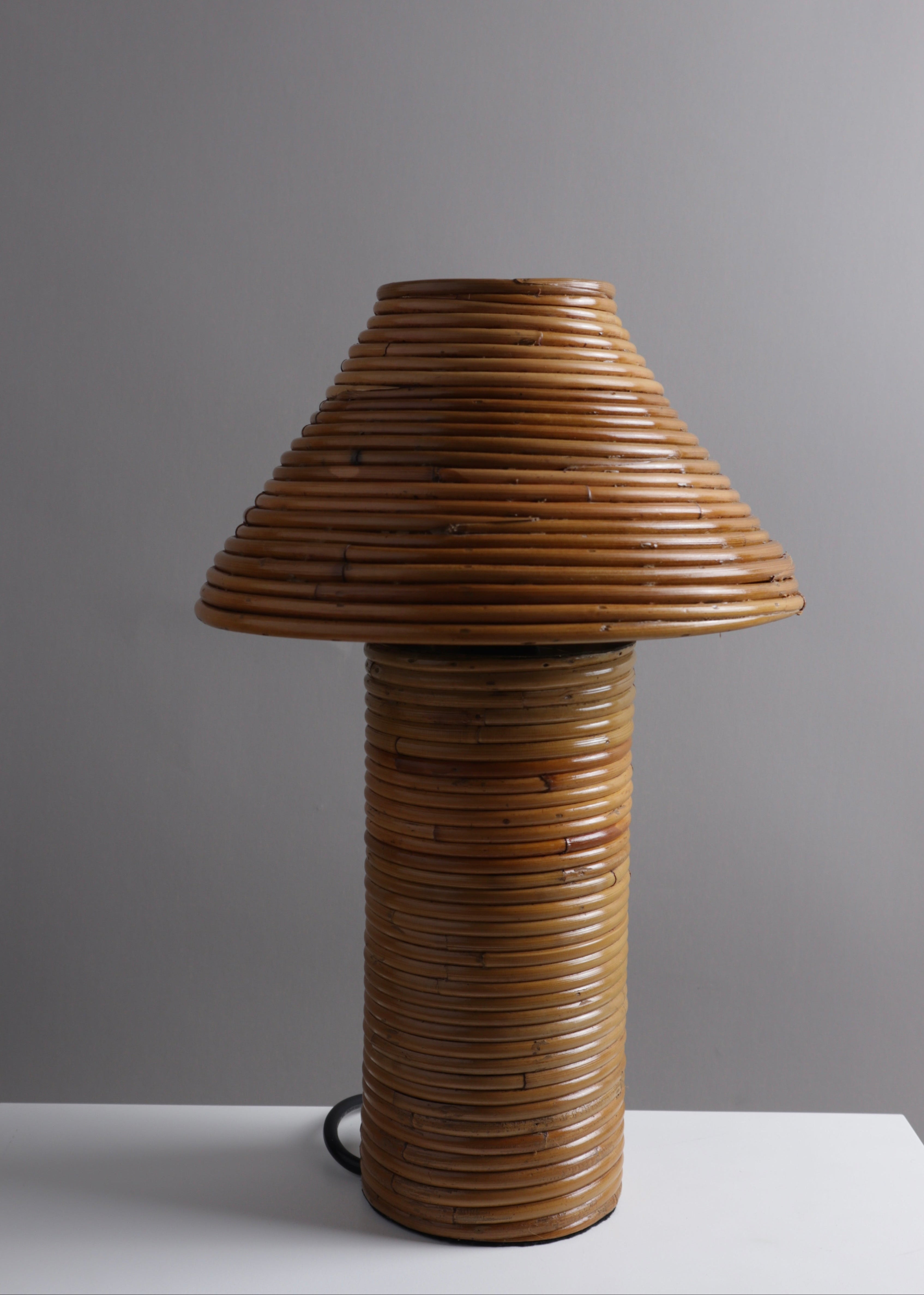 A mid-century modern table lamp made of tightly wrapped natural rattan or bamboo, featuring a conical shade and a cylindrical base, set against a neutral gray background.