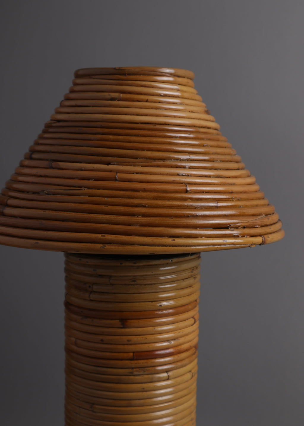 A rustic lamp made from tightly wrapped natural bamboo or rattan, featuring a conical shade and a cylindrical base, set against a solid dark gray background.