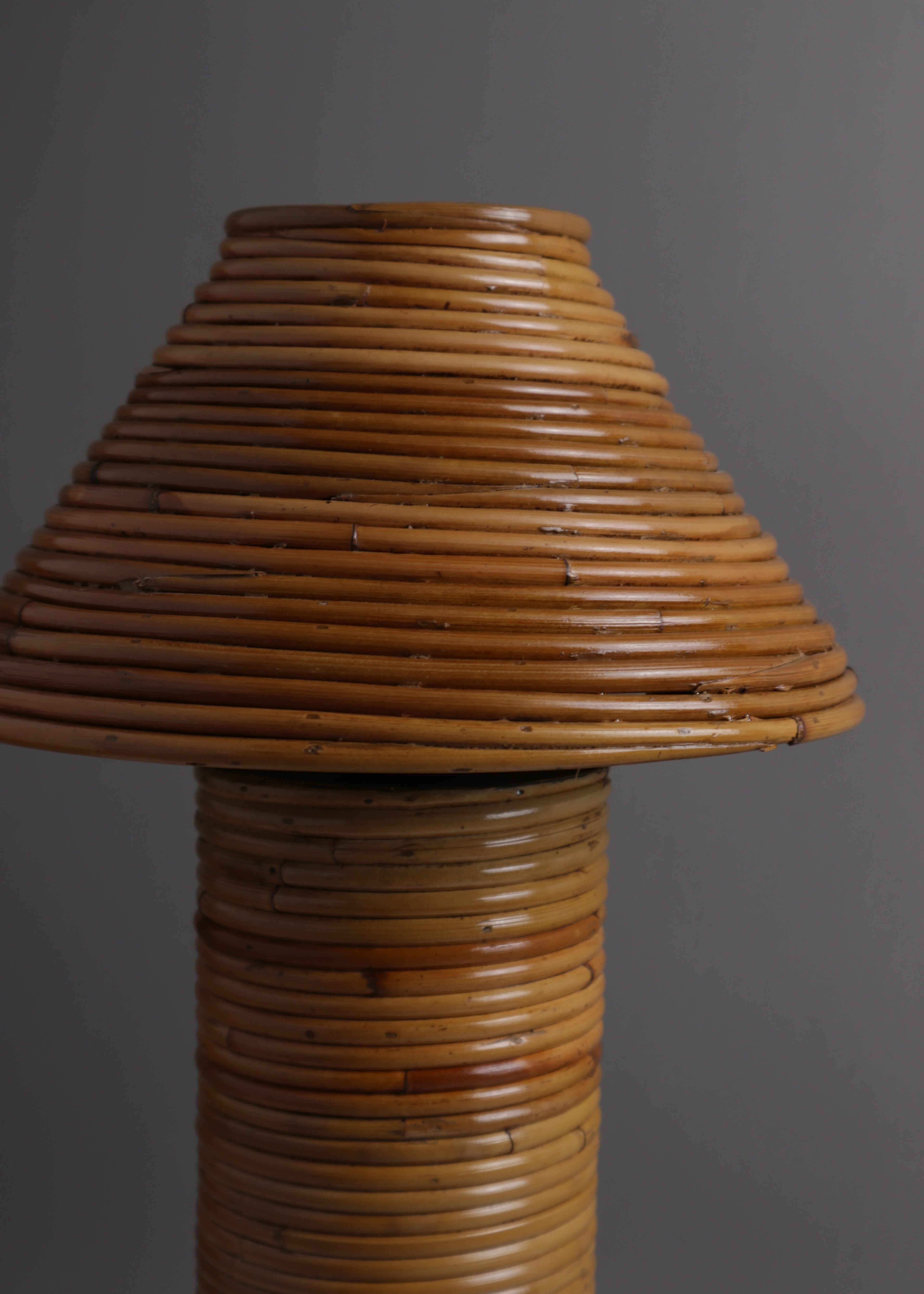 A rustic lamp made from tightly wrapped natural bamboo or rattan, featuring a conical shade and a cylindrical base, set against a solid dark gray background.
